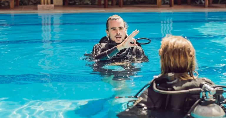 A PADI Basic Scuba Diving Course in Dubai instructor makes a hand signal while teaching a student in a swimming pool, with both individuals wearing full scuba diving equipment.