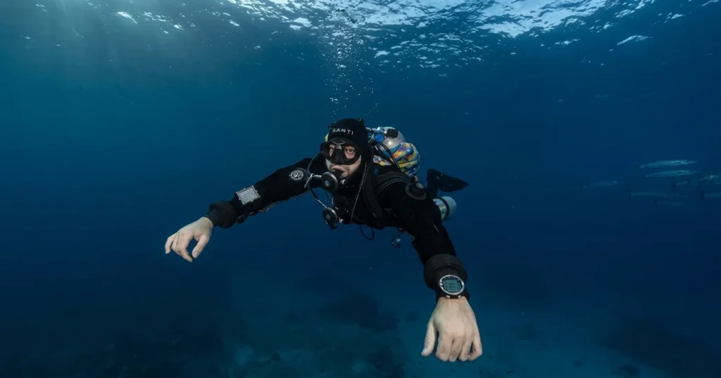 A crisp, high-resolution underwater photograph of a technical scuba diver in full gear with a drysuit, twin back-mounted cylinders, a side-mounted stage tank, and long-hose regulators, swimming in deep blue water. Sunlight caustics are visible from the surface. This image represents the technical skills learned in a Tec Basics Course in Dubai: Building the Foundation Before Tec 40