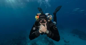 A detailed underwater photograph of a technical scuba diver in a full black SANTI drysuit, twin back-mounted cylinders, and a side-mounted stage tank, hovering over a sandy seabed. The diver looks at the camera, holding a gloved hand near their mouth in a focused pose, demonstrating skills for a Tec Basics Course in Dubai. Sunlight rays penetrate from the surface, and a school of barracuda is visible in the distance